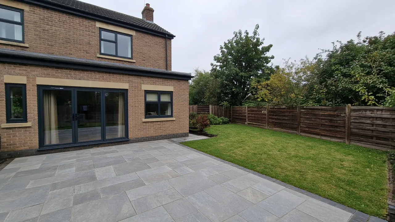 Example image: grey sandstone patio behind a modern detached house with a lawn and fence